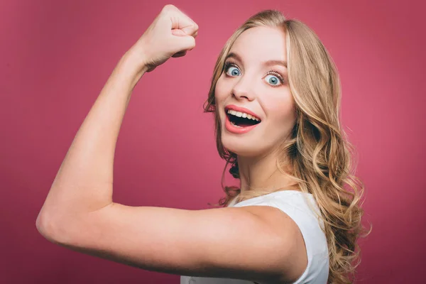 Starkes und kraftvolles Mädchen zeigt Muskeln an ihrer Hand und lächelt. Sie schaut in die Kamera. isoliert auf rosa Hintergrund. — Stockfoto
