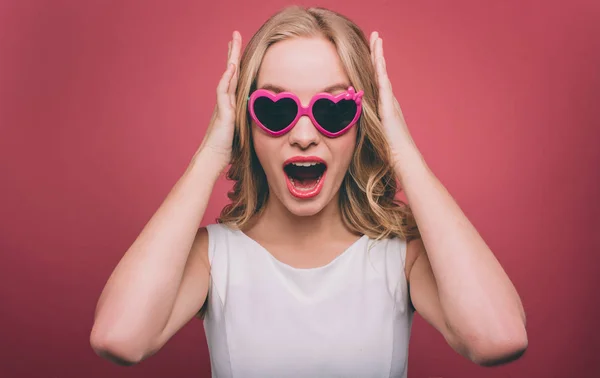 Förvånad och attraktiva flicka bär solglasögon. Hon tittar rakt fram. Kvinnliga person håller händerna på huvudet. Isolerad på rosa bakgrund. — Stockfoto