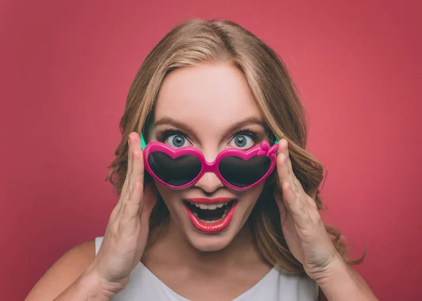 Hübsche Frau mit blonden Haaren trägt Sonnenbrille mit rosa Rand. Sie blickt in die Kamera und hält eine Brille an der Hand. sie glücklich und gut aussehend. isoliert auf rosa Hintergrund. — Stockfoto