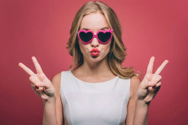 Das Bild eines Mädchens mit blonden Haaren blickt geradeaus durch eine Brille mit rosa Rand. Sie zeigt das Stück sybol mit ihren Fingern und macht ihre Lippen in Kussform. isoliert auf rosa — Stockfoto