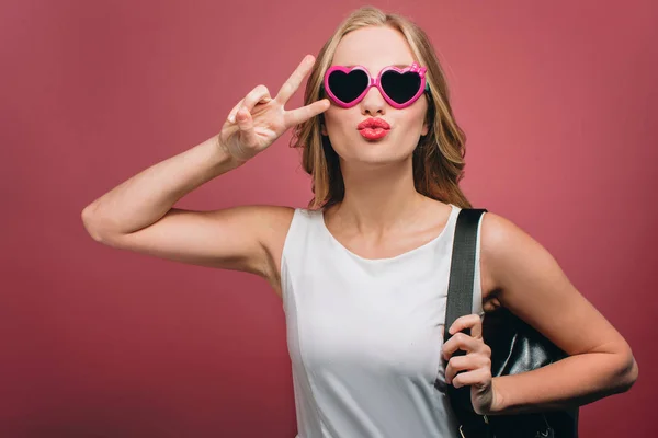 Bella ragazza con gli occhiali e borsa nera è loooking dritto. Sta mostrando il simbolo del pezzo e mettendo le labbra in una forma di bacio. Isolato su sfondo rosa . — Foto Stock