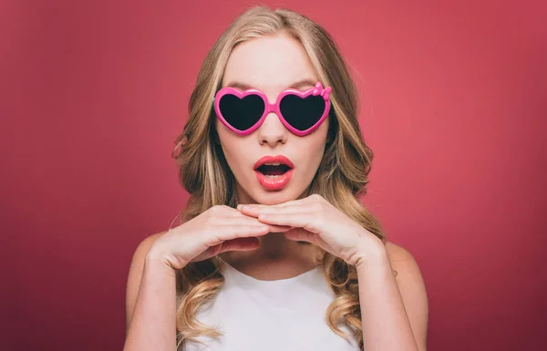 Attraktive und staunende junge Frau posiert. Sie blickt geradeaus durch eine Brille mit rosa Rand in die Kamera. Sie hält ihre Hände unter dem Kinn. isoliert auf rosa Hintergrund. — Stockfoto