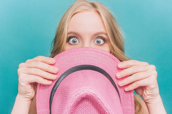 Förvånad och överraskad tjej tittar rakt på kameran med sin stora och blå ögon. Hon täcker en del av ansiktet med rosa hatt. Isolerad på blå bakgrund. — Stockfoto