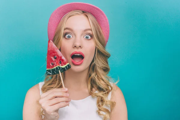 Förvånad och överraskad tjej bär Rosa hatt. Också håller hon lollipop på minnet. Ung kvinna tittar rakt fram. Isolerad på blå bakgrund. — Stockfoto