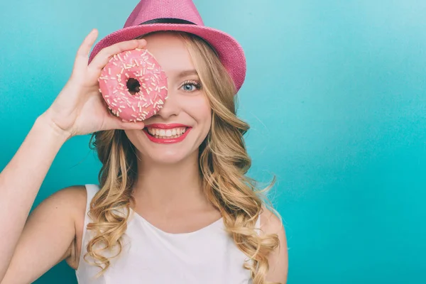 Attraktives Mädchen mit blonden Haaren sieht gerade aus und lächelt. Sie bedeckt eines ihrer Augen mit Donut mit rosa Glasur. Mädchen ist glücklich. isoliert auf blauem Hintergrund. — Stockfoto