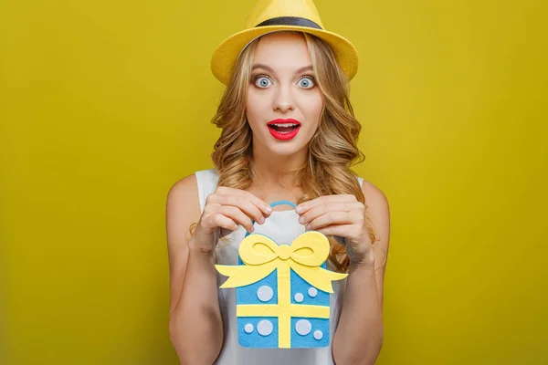 Mycket glada tjej håller en blå med gula gåva i händerna. Hon är förvånad över mycket. Isolerad på gul bakgrund. — Stockfoto