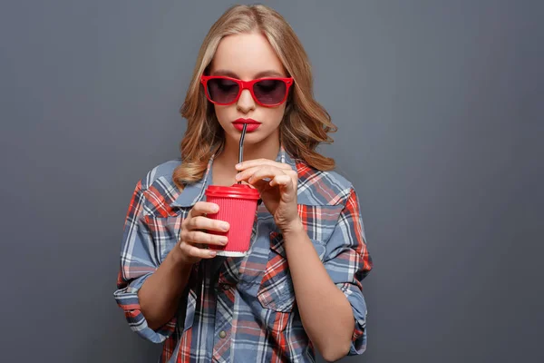 Cool girl wears dark glasses with red edge. She is drinking coke from the red cup. Isolated on grey background.