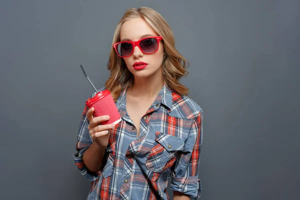 Mädchen mit blonden Haaren trägt dunkle Brille mit rotem Rand. Sie hält eine Tasse Cola in der Hand. Mädchen blickt geradeaus. isoliert auf grauem Hintergrund. — Stockfoto