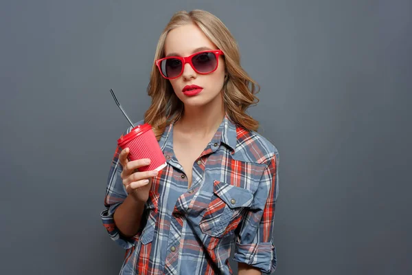 Tjej med blont hår bär mörka glasögon med röd kant. Hon håller en kopp av koks. Flickan tittar rakt fram. Isolerad på grå bakgrund. — Stockfoto