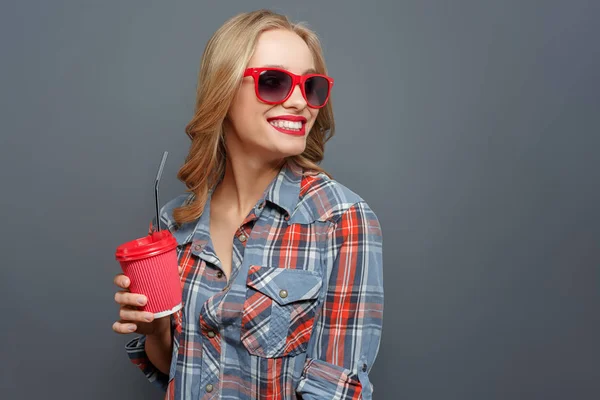 Freundliches Mädchen blickt nach rechts. , lächelt sie. Die junge Frau hält einen roten Becher Cola in der Hand. Sie trägt grau mit rotem Hemd. isoliert auf grauem Hintergrund. — Stockfoto
