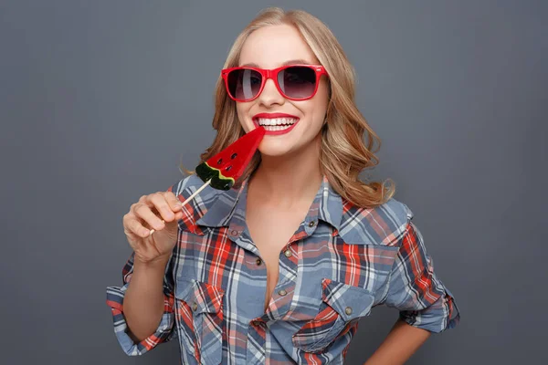 Divertente foto di ragazza che morde il lecca-lecca. Sta sorridendo. Anche questa signora indossa occhiali scuri con bordo rosso. Isolato su sfondo grigio . — Foto Stock