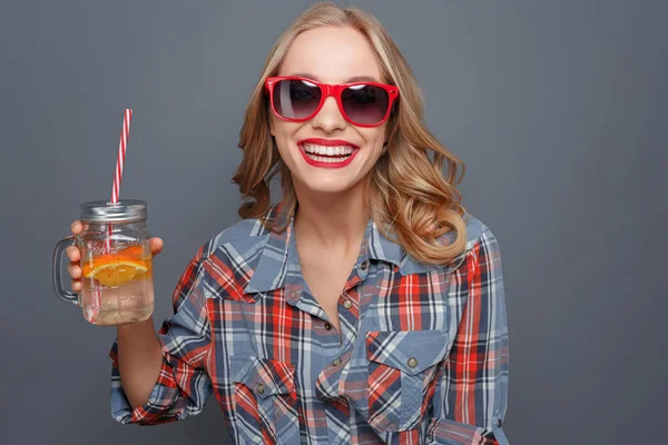 Awesome och trevlig tjej håller en kopp med lite juice och en bit av orange i den. Hon skrattar. Flickan bär mörka glasögon med röd kant. Isolerad på grå bakgrund. — Stockfoto