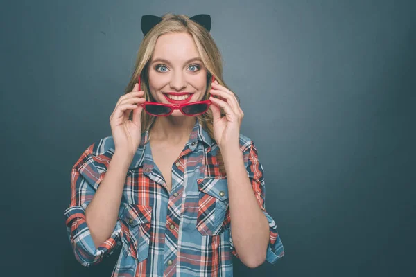 Niza y feliz persona está usando gris con camisa roja y una oreja de gatito en la cabeza. También se está poniendo las gafas y sonriendo. La chica está mirando hacia adelante. Aislado sobre fondo azul . — Foto de Stock