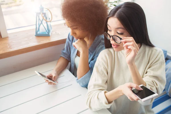 Am Tisch sitzen zwei beste Freundinnen. Afroamerikanerin liest am Telefon, während ihr Freund mit einer Hand die Brille hält und zum Handy ihrer Freunde schaut. — Stockfoto