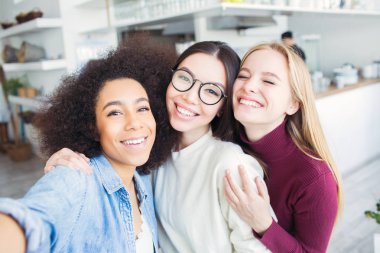 Üç güzel arkadaş birlikte selfie alıyor. Orada afro Amerikalı kız, bir esmer ve bir sarışın. Bunlar kameraya bakıyor ve gülümsüyor. Kızlar çok neşeli.