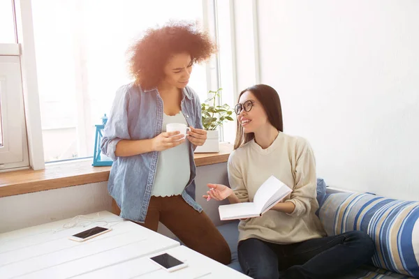 Friends are gathered together in a room. One of them is standing with a cup of tea and looking to the notebook while the other one is sitting and holding a notebook. They are smiling.