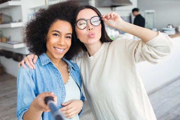 En annan bild av två vänner. De gör selfie. Flicka i blå skjorta hålla en selfie-stick och leende kameran medan hennes vänner är poserar och skicka kyssar till kamera. — Stockfoto