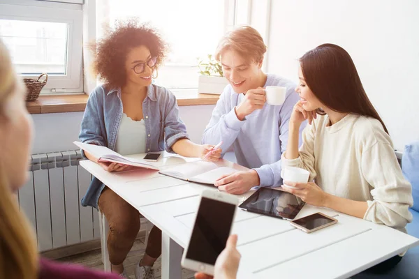 Vier Personen sitzen am Tisch und studieren. Mann und Mädchen im weißen Pullover schauen zum Notizbuch, während Mädchen im blauen Hemd ihnen einen Stift reichen. ein anderer schaut auf den Bildschirm der Telefone. — Stockfoto