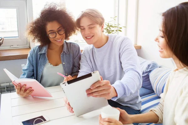 Nettes Bild von Kerl, der einem Mädchen im blauen Hemd etwas auf dem Tablet zeigt. ihr Freund im weißen Pullover sitzt neben ihnen und hält eine Tasse Tee. — Stockfoto