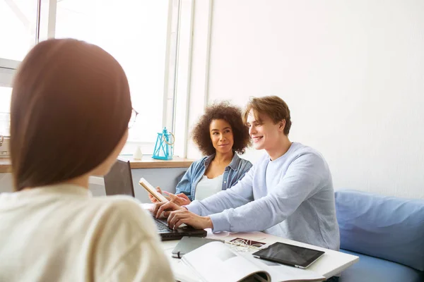 Afroamerikanerin und ein Gey sitzen zusammen auf dem Sofa. Er zeigt ihr etwas auf dem Laptop. Sie schaut ihn an. Ein anderes Mädchen sitzt vor ihm. — Stockfoto