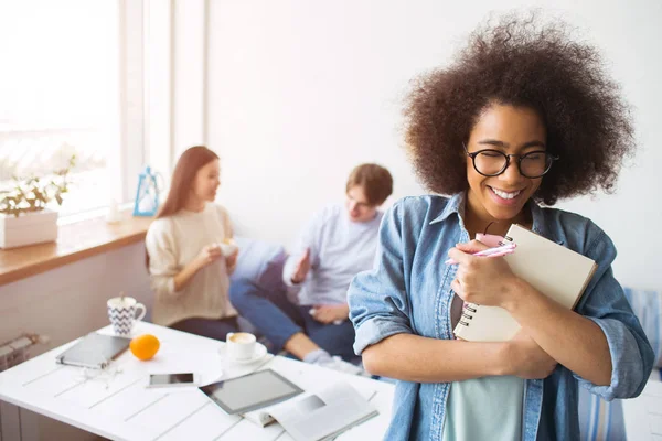 Afroamerikanisches Mädchen steht vor der Kamera und hält ein Notizbuch. sie lächelt und kichert. ihre Freundinnen sitzen auf dem Sofa hinter ihr und reden. — Stockfoto