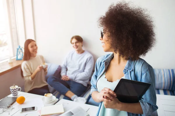 Afroamerikanerin hält ein Tablet in der Hand und schaut ihre Freundinnen an, die auf dem Sofa sitzen, lächeln und sie anschauen. — Stockfoto