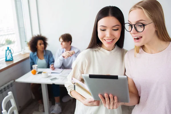 Twee slimme meisjes zijn permanent en kijken op tablet. Het blonde meisje draagt bril. Haar vrienden is het bezit van een laptop. Er zijn hun vrienden zitten achter hen en studeren. — Stockfoto