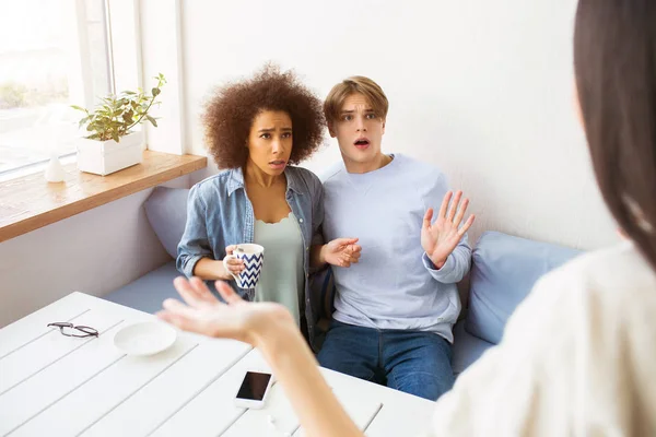 Mädchen im weißen Pullover steht vor ihren Freundinnen und winkt mit der Hand. Ein afroamerikanisches Mädchen und ein Mann liegen auf dem Sofa. sie sehen verwirrt und unglücklich aus. — Stockfoto
