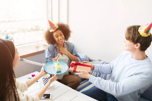 Glada vänner firar födelsedag. De har roliga hattar på huvudet. Killen håller en present medan flickan i vit tröja håller plattan med kaka. Afro amerikansk flicka är glad. — Stockfoto