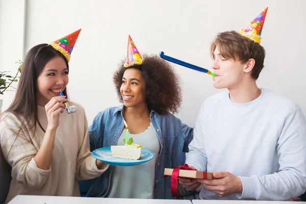Drei Menschen feiern Geburtstag. Sie tragen lustige Hüte. Mädchen hält einen Teller mit Kuchen, während Kerl ein Geschenk in der Hand und ein seltsames Ding im Mund hat. — Stockfoto