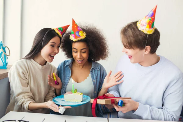 Drei Menschen feiern Geburtstag. Sie tragen lustige Hüte. Mädchen hält einen Teller mit Kuchen, während Kerl ein Geschenk in der Hand und ein seltsames Ding im Mund hat. — Stockfoto