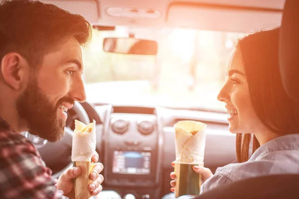 Schöne Menschen sitzen zusammen im Auto und schauen einander zu. Sie halten Hühnerbrötchen in der Hand und lächeln. Auto steht im Wald. — Stockfoto