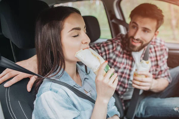 Girl is eating chicken roll with pleasure. She is enjoying the moment. Guy is holding the same roll in his hands and looking at her with excitement. They have their seatbelts on.