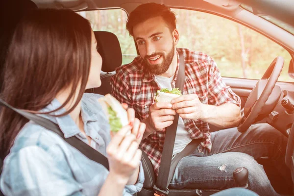 Joli et beau couple est assis ensemble dans la voiture et regardant vers l'autre. Ils sont verrouillés avec la ceinture de sécurité. Les gens tiennent des sandwichs. Ils se sont arrêtés pour manger. . — Photo