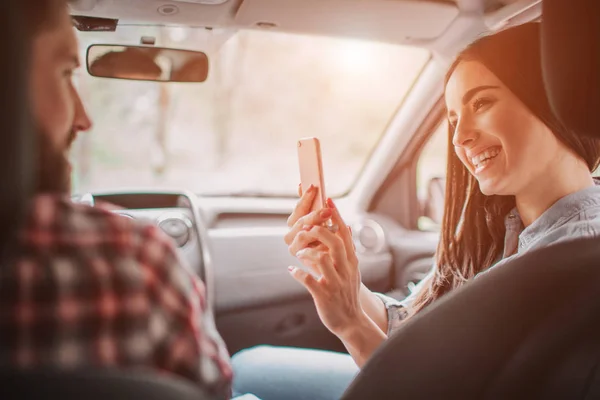 La joven está tomando fotos de su hombre. Ella está mirando el teléfono y sonriendo. Ella está haciendo eso en el coche . — Foto de Stock