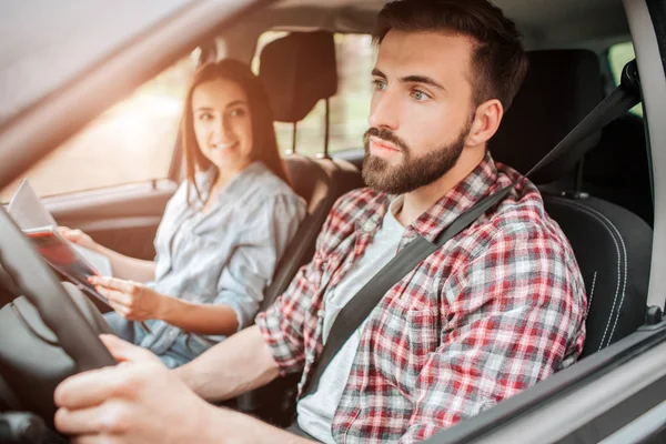 Cuidado y buen tipo está conduciendo el coche y mirando hacia adelante. Está prestando toda su atención al camino. La chica está sentada a su lado. Ella sostiene el mapa y mira al joven con sonrisa . — Foto de Stock