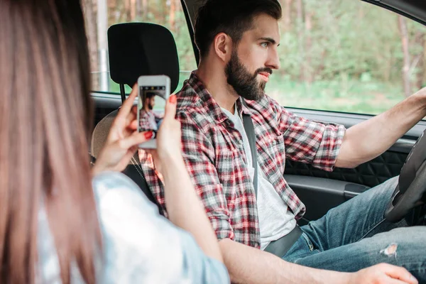 La fille prend une photo de son beau petit ami. Il ne regarde pas la caméra mais pose et conduit une voiture . — Photo