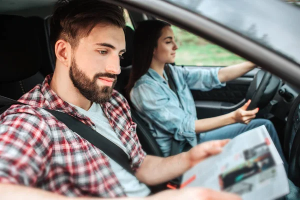 Nice people are riding in car. Girl is driving machine. Guy is sitting besides her and reading a book. They are travelling and spending time together.