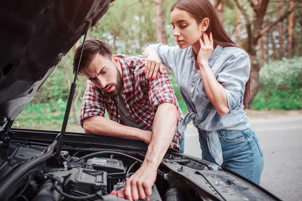 Un tipo inteligente y serio está arreglando el coche. Chica está de pie a su lado y mirando al proceso. Ella se apoya en su novio . — Foto de Stock