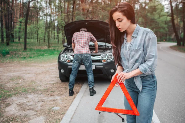 Dziewczyna trzyma czerwony trójkąt na ziemię. Ona jest właśnie najlepsze miejsce, aby to zrobić. Facet jest stojący przed czarnym samochodem i patrząc pod kaptur. — Zdjęcie stockowe