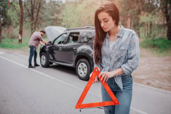 Ładne i poważne dziewczyna jest stojący na drodze i posiadający znak pf czerwony trójkąt. Patrzy w dół. Facet jest stały oprócz samochodów i mocowania. — Zdjęcie stockowe