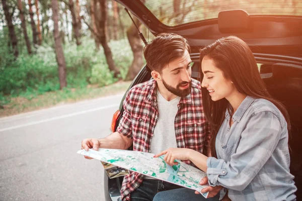 Beutiful people are sitting in trunk and talking to each other. Guy is holding a map and looking at girl while she is pointing on map and smiling. She is studying it. — Stock Photo, Image