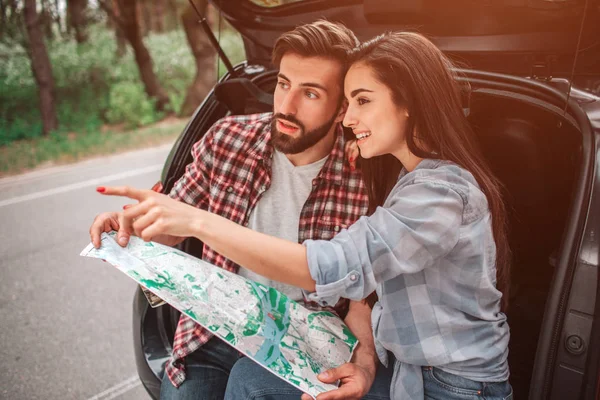 Attractive girl is sitting with her boyfriend in cars trunk and pointing straight forward. She is smiling. Guy is holding a map and looking straight too. — Stock Photo, Image