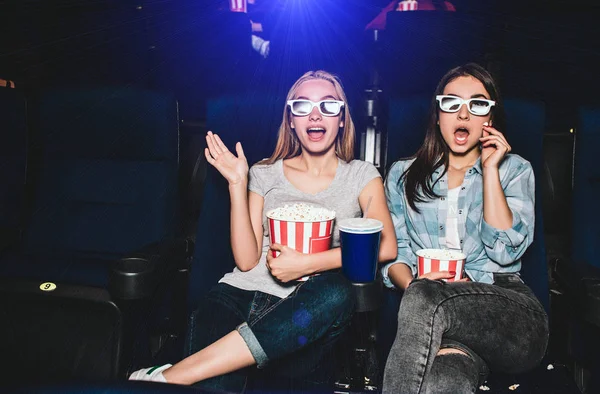 Scared and excited girls are sitting in a cinema hall and watching movie. They are amazed. Also girls have baskets of popcorn and a big cup of cola. — Stock Photo, Image