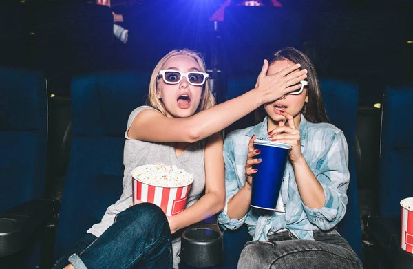 Girls are terrified. They are watching a horror movie. Blonde girl is covering eyes of her friend. Brunette is trying to drink some cola from big cup. — Stock Photo, Image