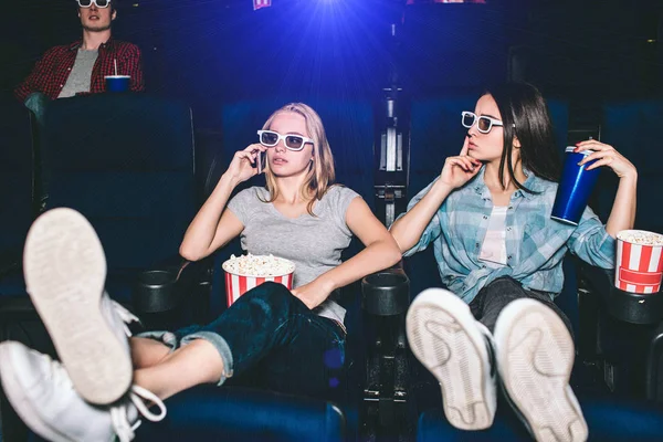Beautiful and attractive girls are sitting in chairs. Blonde girl is talking on thhe phone. Her friend is showing the silence symbol. She wants her to be quiet and stop talking during watching movie. — Stock Photo, Image