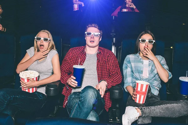 Scared and frightened people are watching movie in cinema. They are sitting and looking straight forward. Everyone has a basket of popcorn or a cup with coke. They are not alone in the hall. — Stock Photo, Image