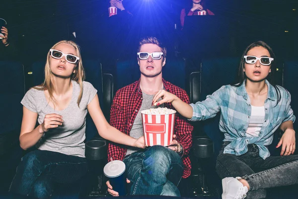 Интересные люди смотрят кино в кино. Они сидят и смотрят прямо сквозь специальные очки. Парень и блондинка держат коробку с попкорном, пока брюнетка ест попкорн . — стоковое фото
