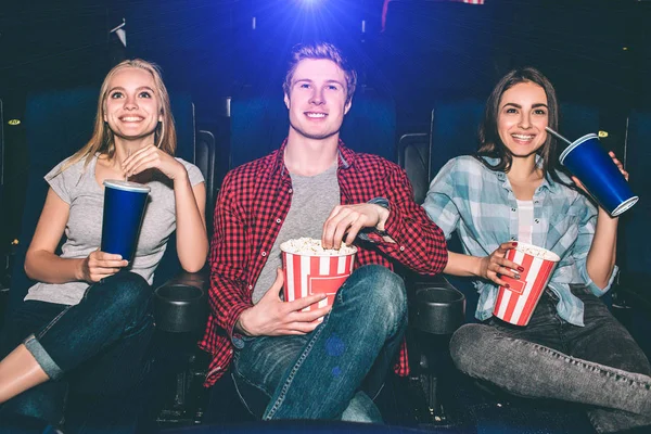 Happy and cheerful people are sitting and watching movie. They are excited. Blonde girl has a cup of cola. Guy holds a basket with popcorn. Brunette girl has all of it in her hands and smiling. — Stock Photo, Image