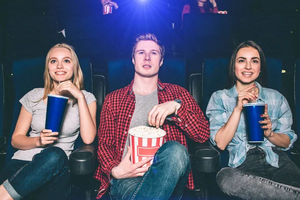 Three beautiful people are sitting in chairs and watching movie. They look happy and excited. Girls are drinking cola and smiling. Guy is very serious. He is holding a basket with popcorn. — Stock Photo, Image